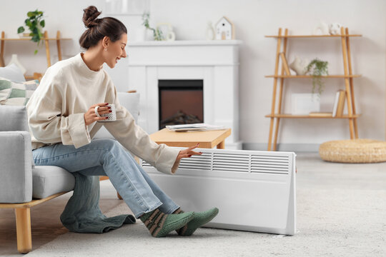 Young woman with cup of tea and electric convector heater sitting on sofa at home