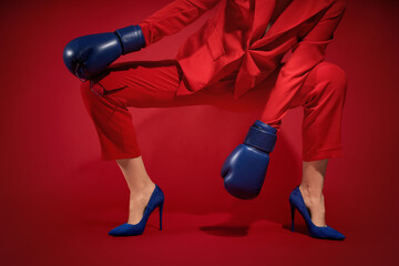 Woman in stylish suit with high heels and boxing gloves on red background