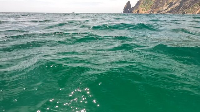 Ocean waves emerald rippling across the deep green sea surface with rocky coastal cliffs in the distance under a bright sky