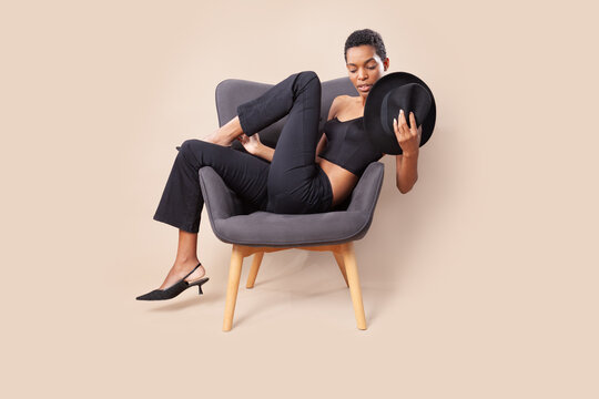 Playful black woman with short hair posing with fedora hat on armchair in photography studio