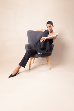 Playful black woman with short hair posing with fedora hat on armchair in photography studio