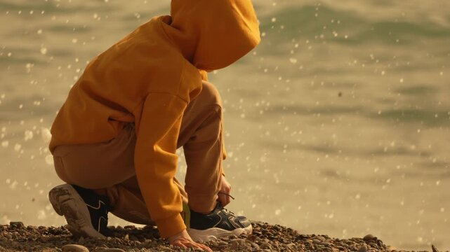 Child beach shoes Young boy in a yellow hoodie crouching on a pebble shore to tie his laces while sea waves splash