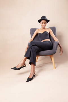 Playful black woman with short hair posing with fedora hat on armchair in photography studio