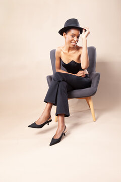 Playful black woman with short hair posing with fedora hat on armchair in photography studio