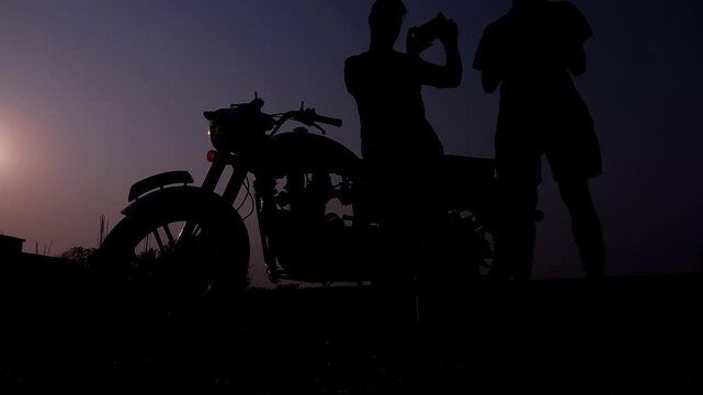 Silhouette of a motorcycle rider standing beside bike during sunset in India. 