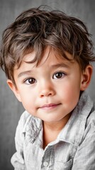 Fototapeta premium Young boy with bright brown eyes and curly hair looks directly at the camera. He wears a light gray collared shirt, posing sweetly in a studio setting against a simple gray background during daytime