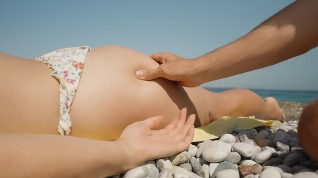 A man's hand squeezes the soft buttocks of a young woman sunbathing on a pebble beach.