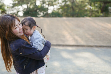 Naklejka premium Mother holds daughter close, smiling gently. Child rests head on her shoulder, eyes closed. Soft sunlight filters through green trees behind them. Open space stretches beyond, calm and quiet