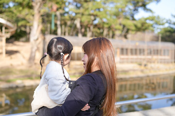 Naklejka premium Mother holds child near calm lake. Sunlight glows softly on both figures. Trees line distant shore behind them. Child gazes away with quiet curiosity. Mother smiles gently, embracing close