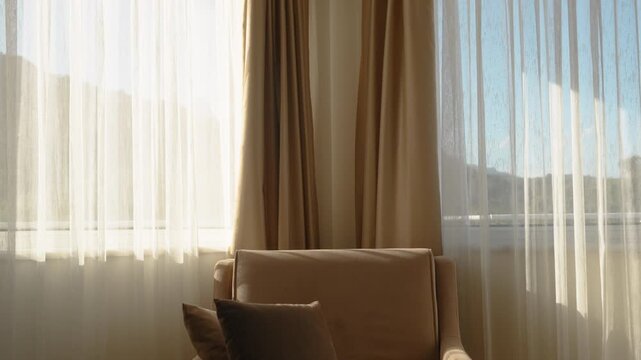 A brown armchair in the room beside windows through which sunlight streams. Curtains diffuse the light.