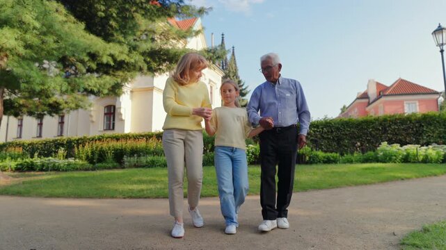 Senior people grandparents walking with granddaughter sunlit park path smiling holding hands urban garden backdrop warm golden light tender family bond active aging moment