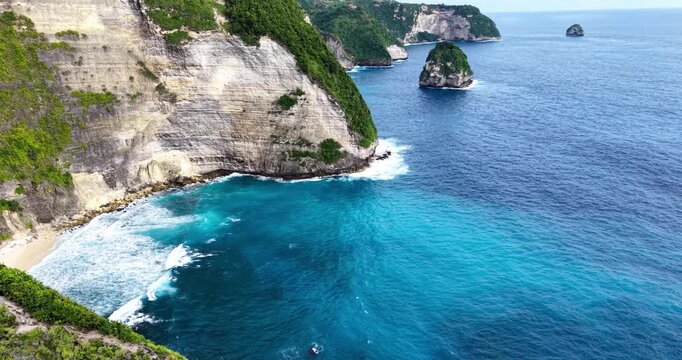 Aerial view of limestone cliffs and turquoise ocean at Kelingking Beach, Nusa Penida, Bali, with waves along a dramatic tropical coastline and offshore rock formation.