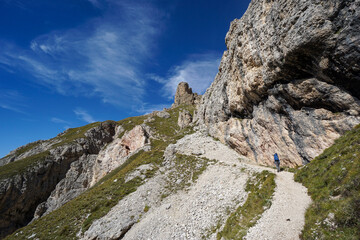 Fototapeta premium Female hiker from behind trekking alpine path with rugged cliffs under clear blue sky