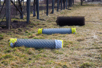 Obraz premium Three rolls of chain-link wire mesh, two new and wrapped, and one old, lay on yellowish grass next to a wooden fence line. Construction materials for fencing, preparation to secure a property in early