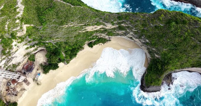 Top-down view of Kelingking Beach, Bali, showing waves meeting a secluded sandy cove beneath steep limestone cliffs. Visible cliffside elevator construction highlights ongoing tourism development.