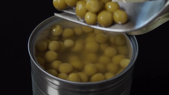 Fork lifting green peas out of open metal can with preserved vegetables in brine