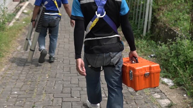 Rear view of a worker in safety harness carrying an orange equipment toolbox on a stone path.