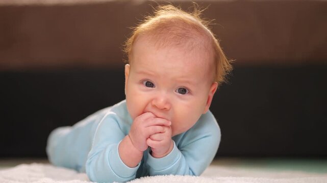 Baby Struggling With Teething Pains. Young Baby In Blue Sleepwear Showing Teething Signs Peacefully. An Adorable Infant Wearing Blue Pajamas Biting Hands With Concentration On Soft Rug Inside