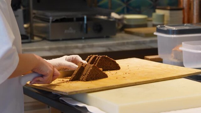Professional Baker Slicing Dark Rye Bread with Seeds. Chef slicing a triangular loaf of dark coriander rye bread on a wooden board. Professional kitchen setting for bakery, restaurant