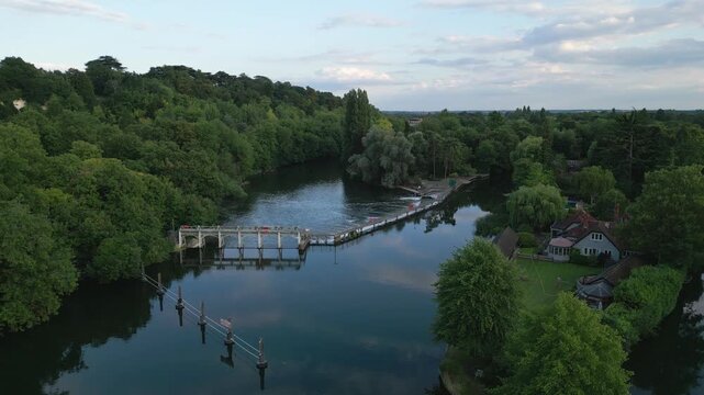 An aerial view of a tranquil river with a weir, bordered by lush green trees and a picturesque house.