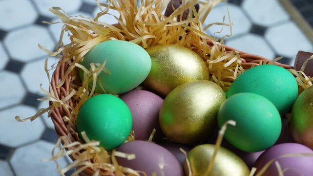 Colorful Easter Eggs in Wicker Basket with Straw. A wicker basket filled with decorative Easter eggs in gold, green, and purple hues nesting on wood wool