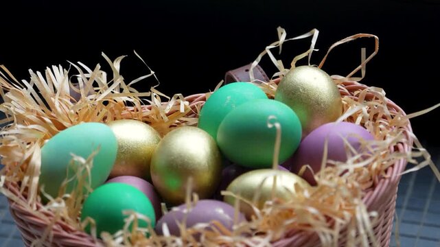 Colorful Easter Eggs in Wicker Basket with Straw. A wicker basket filled with decorative Easter eggs in gold, green, and purple hues nesting on wood wool