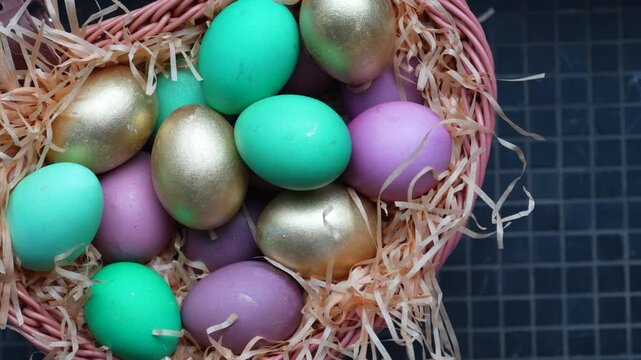 Colorful Easter Eggs in Wicker Basket with Straw. A wicker basket filled with decorative Easter eggs in gold, green, and purple hues nesting on wood wool
