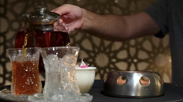 Close-up of Hand Pouring Hot Black Tea into Armudu Glass. A person hand pouring hot tea from a glass teapot into a traditional Azerbaijani armudu glass against a dark, atmospheric background