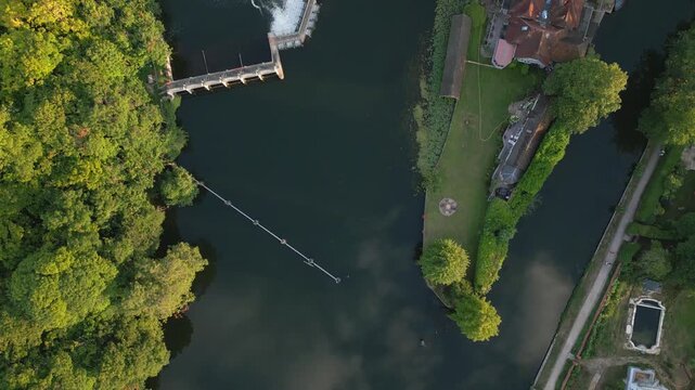 Aerial view of a dam, lush trees, and houses bordering a dark river. Features include a footbridge and a swimming pool.