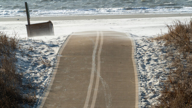 Wind-swept beach access path leading to a turbulent ocean with foamy waves and blowing sand across coastal dunes and dry grass on a blustery day.