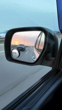 View of a golden sunset and trailing traffic reflected in a vehicle's side-view mirror. Motion blur on the asphalt creates a sense of speed and travel during a scenic evening road trip
