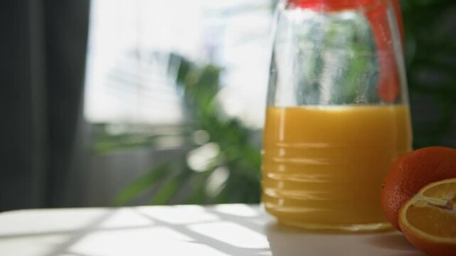 Detailed close-up: male hand takes glass of fresh orange juice on table in bright bedroom. Indoor palm tree in soft focus, warm sunlight pours through window, healthy vitamin breakfast moment 