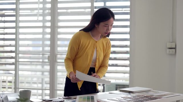 Asian adult female designer reviewing architectural plans color palettes project proposals in modern bright office studio professional creative workspace