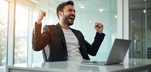 Naklejka premium Man in black blazer cheers with joy at laptop. He celebrates success in modern office. Bright workspace with glass walls provides natural light. He shows winning emotion.