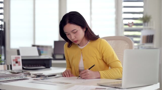 Asian adult woman drawing design artwork focused working modern office workspace creative professional planning project inspiration color palette desk laptop