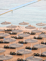 Fototapeta premium Aerial view of rows of straw beach umbrellas and sun loungers on a sandy beach at a luxury resort in Sharm El Sheikh, Egypt, bordering the clear turquoise waters of the Red Sea.
