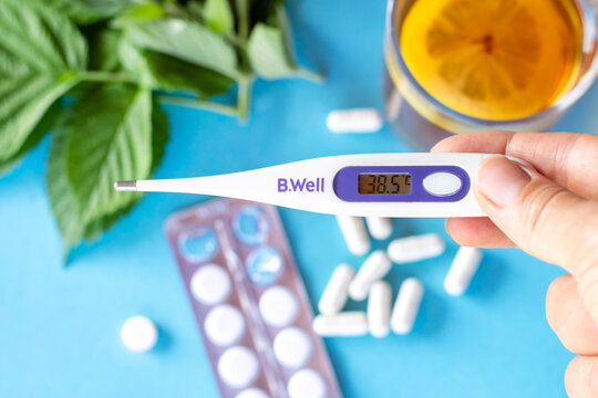 A thermometer showing a temperature of 38.5 degrees in the hand against the background of tea with lemon and tablets (medicines). The concept of seasonal diseases, the treatment of diseases