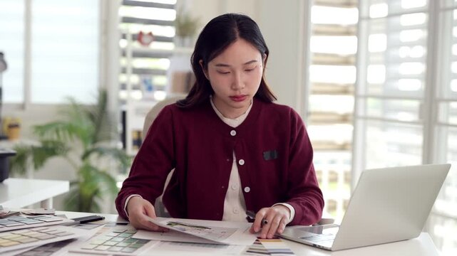Asian adult woman professional architect interior designer working on design plans color swatches laptop modern office studio creative project workspace
