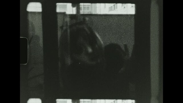 A spooky old doll face peers out in black and white from a dark balcony against an urban background on vintage super 8 film scan