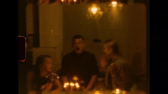 A happy family of three smiles around a table with lit birthday candles under warm lamps on super 8 film scan