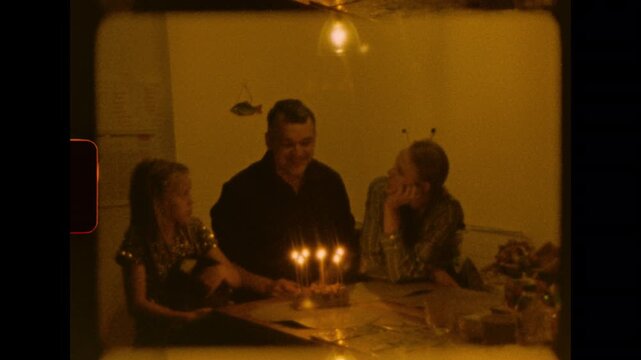 A family is gathered around a table with a lit birthday cake in a cozy room at night on analog super 8 film