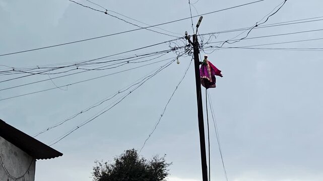 A Gudi, or Gudhi Padwa flag, is seen caught on electrical wires atop a tall pole. The celebratory item, featuring a pink cloth and metal pot, poses a significant risk of electrocution or causing a sho