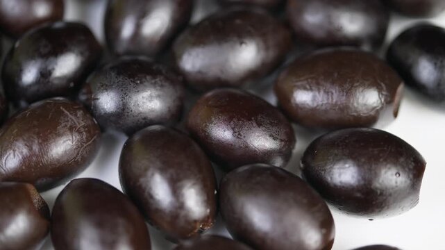 Studio shot of textured black olives. Photograph highlighting oily wrinkled olives against white surface. Detailed studio image showcasing glossy black olives with textured skins