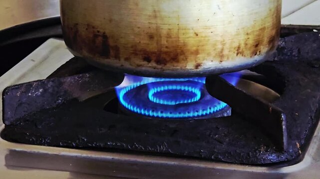 A pot sits over a blue gas flame on a grimy, worn stove. This simple act of cooking highlights the global gas crisis, where price hike & supply shortages turn basic energy needs into a costly struggle
