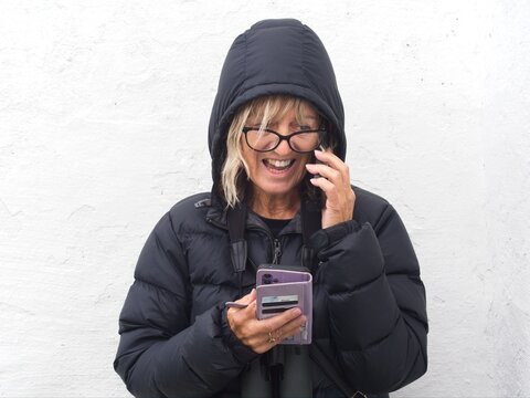 A lady with hood up speaks on a phone and is seen laughing.She holds another phone in the other hand and has binoculars around her neck
