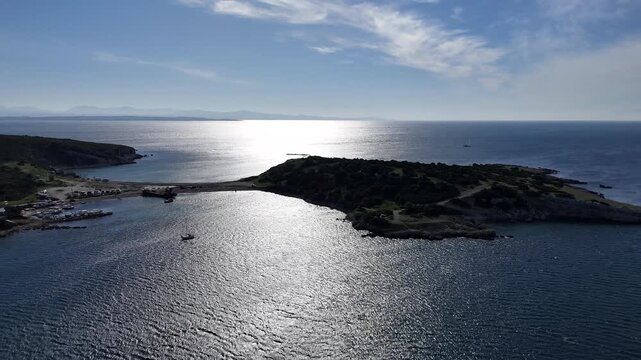 A high-altitude aerial view showcasing a rugged Mediterranean island connected to a mainland bay by a narrow, sandy tombolo.
