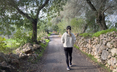Fototapeta premium A young woman seen from behind walking along a forest path, surrounded by trees and greenery. Concept of solitude, harmony with nature, outdoor lifestyle, and peaceful exploration.
