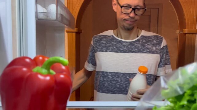 Man takes milk and items from fridge in kitchen with fresh vegetables