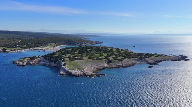 A high-altitude aerial view showcasing a rugged Mediterranean island connected to a mainland bay by a narrow, sandy tombolo.