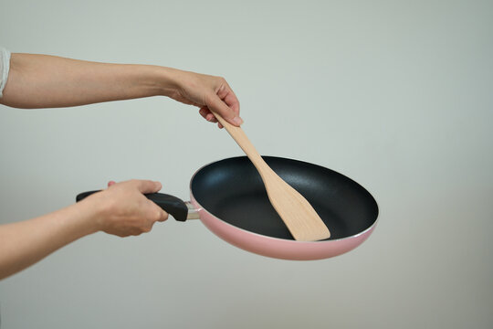 Frying pan in human hands, Cooking concept. Two hands hold a pink frying pan and wooden spatula against a light background, showcasing kitchen utensils.
